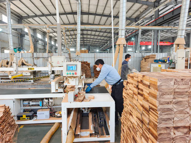 A worker handling small acacia pieces at a sanding station beside stacked blanks